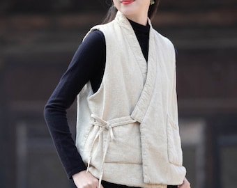 May include: A woman wearing a light beige linen vest with a tie closure. The vest has a relaxed fit and a slightly asymmetrical hem. The woman is also wearing a black turtleneck shirt and black pants.
