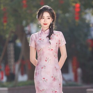 May include: A woman wearing a pink and white floral print cheongsam dress with a high collar and short sleeves. The dress has a side slit and a tassel detail. The woman has her hair pulled back in a braid and is wearing pearl earrings.