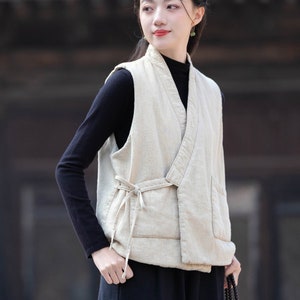 May include: A woman wearing a light beige linen vest with a tie closure. The vest has a relaxed fit and a slightly asymmetrical hem. The woman is also wearing a black turtleneck shirt and black pants.