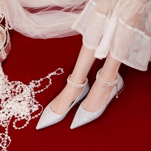 May include: A pair of silver sparkly pointed-toe flats with pearl ankle straps. The shoes are on a red surface.