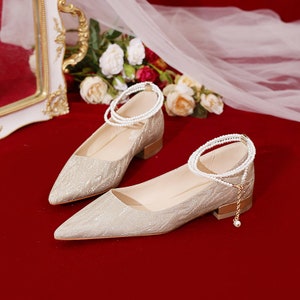 May include: A pair of gold sparkly pointed-toe flats with a low heel. Each shoe has a pearl ankle strap.