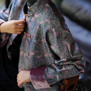 May include: A patterned jacket in shades of green and pink with a mandarin collar and gold-colored buttons. The jacket has long sleeves with a velvet cuff. The fabric features an intricate design.