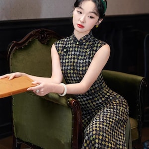 May include: A sleeveless black and gold chequered pattern dress with a high collar and side slit. The dress is worn with a green headband and white bracelet. The person is seated in a green velvet chair.