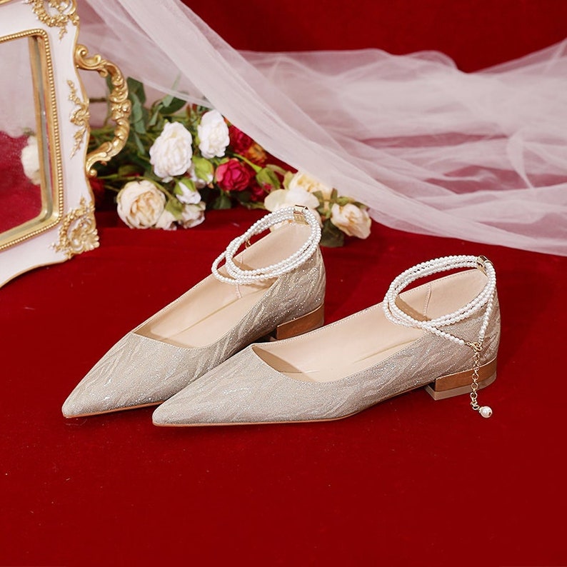 May include: A pair of silver sparkly pointed-toe flats with a low heel. Each shoe has a pearl ankle strap.
