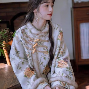 May include: A cream-coloured floral jacket with a mandarin collar and toggle closures. The jacket features a botanical print with yellow, orange, and purple flowers. The sleeves have fur trim at the cuffs. The jacket is part of a traditional clothing collection.