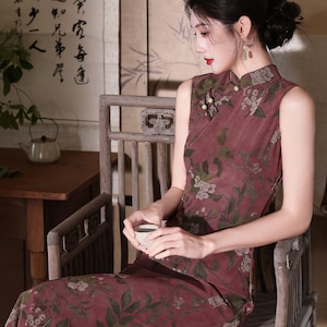 May include: A sleeveless, floor-length dress in a muted mauve-red hue featuring a dark green and white floral botanical print. The garment has a high mandarin collar with decorative fabric knot buttons along the side and a side slit hemline.