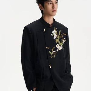 Puede incluir: Chaqueta negra con cuello mandarín y bordado floral. La chaqueta presenta un cierre de botón único y un corte entallado. El bordado incluye flores blancas y hojas verdes, añadiendo un toque de elegancia a la prenda.
