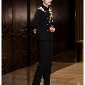May include: A black suit with gold embroidery on the shoulders and chest. The suit includes a mandarin collar jacket and matching trousers. The person is wearing glasses and black dress shoes. The background is a dark wood panel.