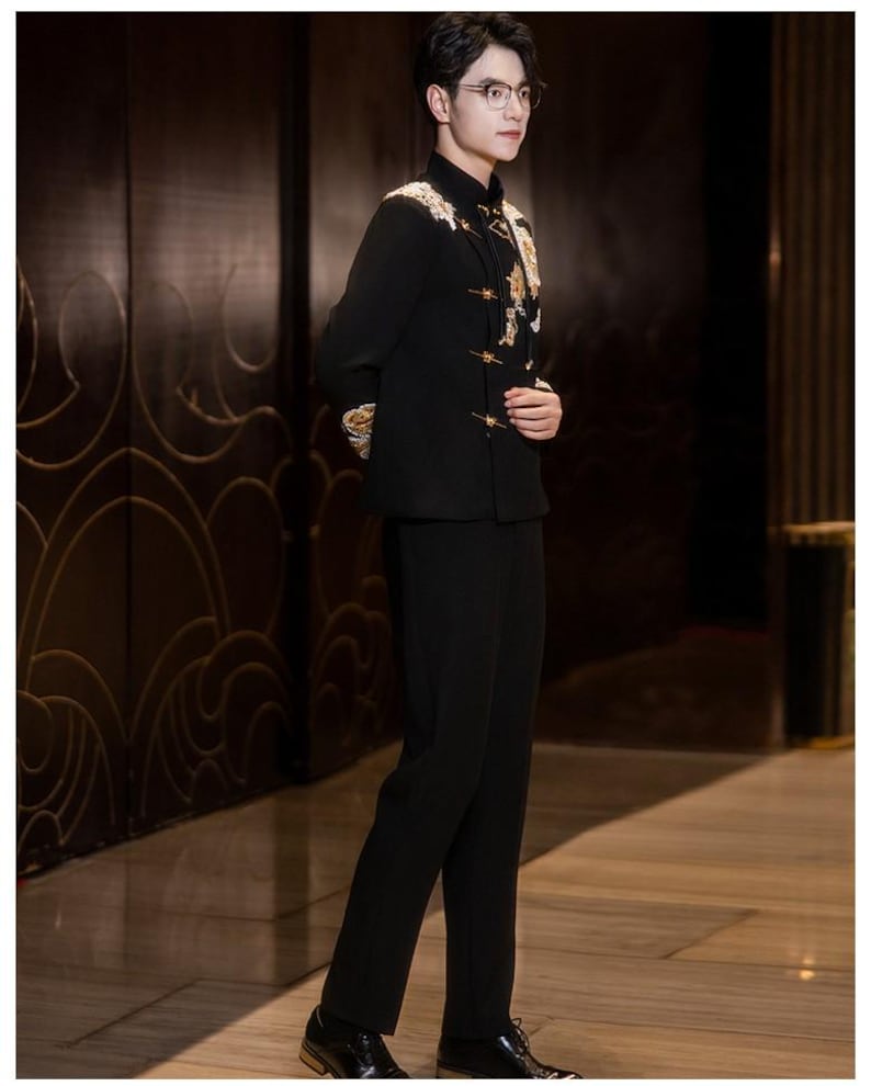 May include: A black suit with gold embroidery on the shoulders and chest. The suit includes a mandarin collar jacket and matching trousers. The person is wearing glasses and black dress shoes. The background is a dark wood panel.