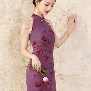 May include: A purple halter dress with a floral pattern. The dress features a high neckline, a small floral brooch, and a side slit. The model is holding a pink tulip. The dress is a formal style.