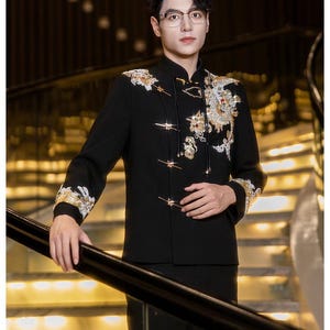 May include: Black formal jacket with gold and white embroidery on the shoulders and sleeves. The jacket has a mandarin collar and decorative frog closures. The sleeves are long and the jacket is worn with black pants.