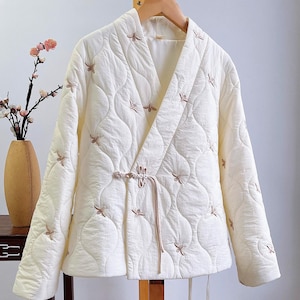 Könnte beinhalten: Cremefarbene Steppjacke mit Wickelverschluss und gestickten Blumenakzenten. Die Jacke hat einen V-Ausschnitt und einen Kordelverschluss. Die Jacke hängt an einem Holzkleiderbügel. Die Jacke ist ca. 71 cm lang.