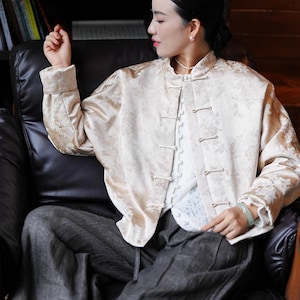 May include: A cream-colored, floral-patterned jacket with a mandarin collar and frog closures. The jacket is worn over a white lace top and paired with gray trousers. The outfit is completed with a jade bracelet.