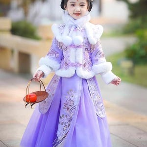 May include: A young girl in a traditional purple and white outfit with fur trim and floral embroidery. She wears a matching headpiece with bunny ears and carries a small basket with a red fruit.