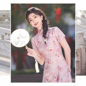 May include: A woman wearing a pink dress with a floral pattern and a white tassel. She is holding a round fan with a floral design. The fan is made of wood and has a white handle.
