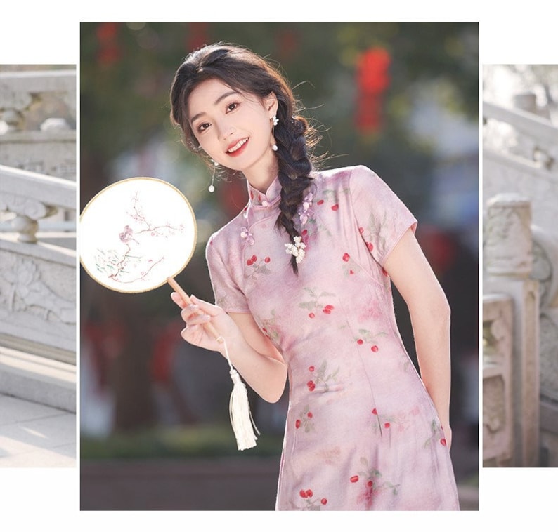 May include: A woman wearing a pink dress with a floral pattern and a white tassel. She is holding a round fan with a floral design. The fan is made of wood and has a white handle.