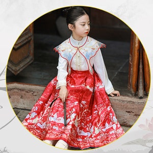 May include: A young girl wearing a white blouse with a red and white embroidered collar and a red skirt with a white floral pattern.