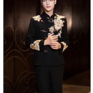 May include: A black suit with ornate gold and white embroidery on the jacket. The jacket has a mandarin collar and frog closures. The person in the image is wearing glasses and has dark hair. The suit is a formal style.