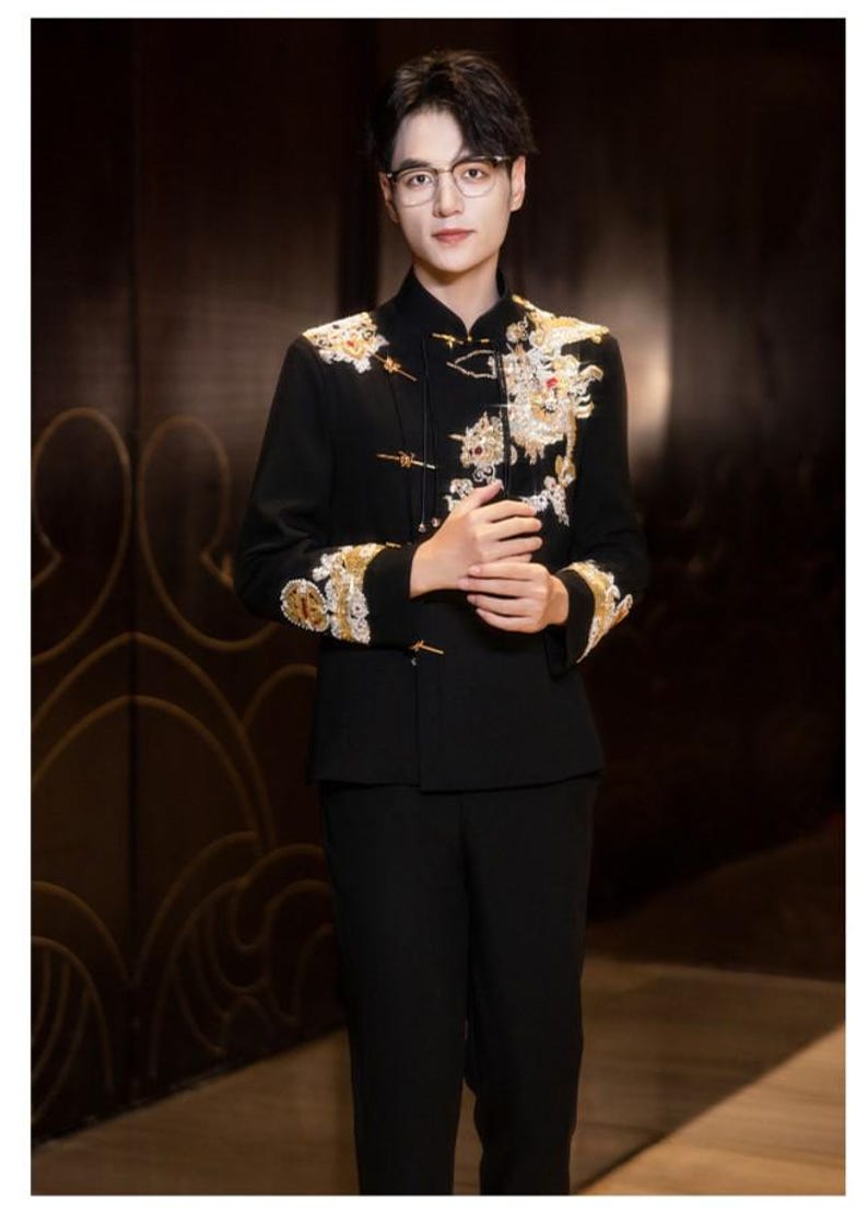 May include: A black suit with ornate gold and white embroidery on the jacket. The jacket has a mandarin collar and frog closures. The person in the image is wearing glasses and has dark hair. The suit is a formal style.