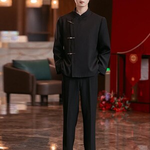 May include: A black suit with a mandarin collar jacket and matching trousers. The jacket features three decorative clasps. The outfit is worn by a person with glasses, standing in a well-lit interior with a polished floor.