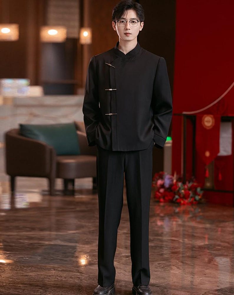 May include: A black suit with a mandarin collar jacket and matching trousers. The jacket features three decorative clasps. The outfit is worn by a person with glasses, standing in a well-lit interior with a polished floor.