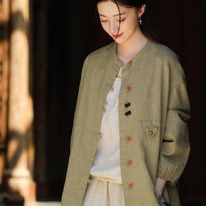 May include: A light olive green linen jacket with a mandarin collar and orange and black buttons. The jacket has two small pockets and is worn over a white top and beige pants. The outfit is a casual, relaxed style.