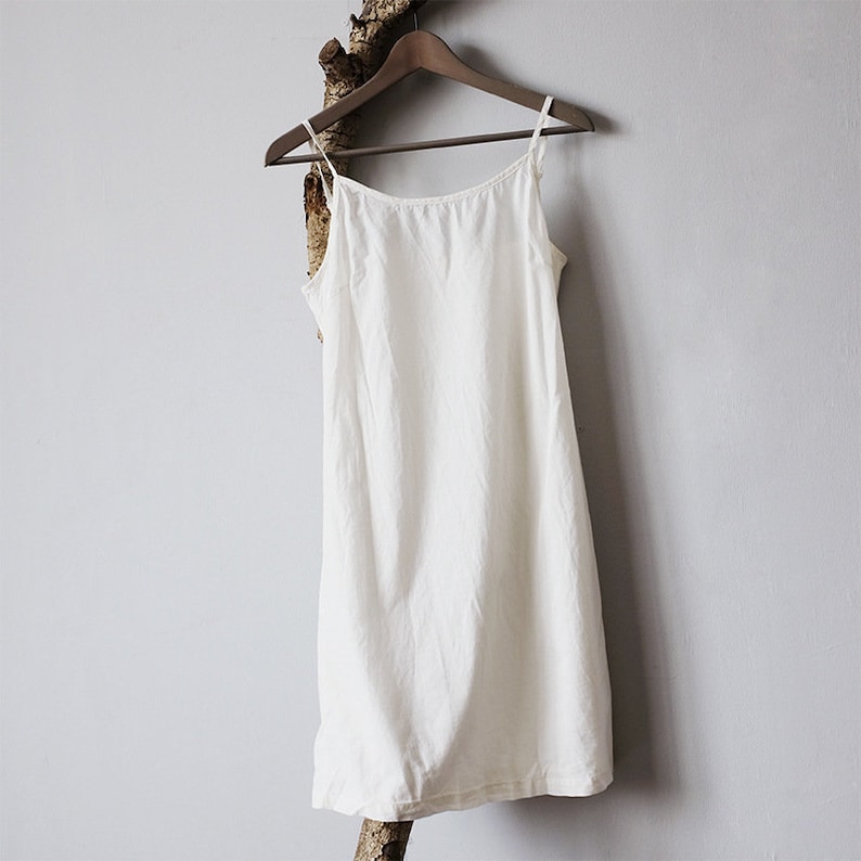 K&ouml;nnte beinhalten: Ein wei&szlig;es, &auml;rmelloses Kleid mit schlichtem Design, das an einem Holzb&uuml;gel an einer wei&szlig;en Wand h&auml;ngt.