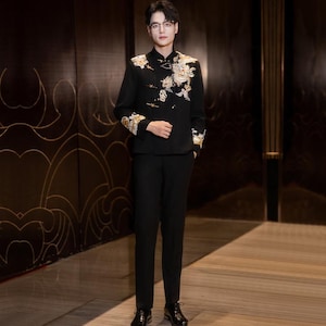 May include: A black suit with gold and white floral embroidery on the jacket. The suit includes a mandarin collar jacket and matching black pants. The jacket's sleeves and shoulders are adorned with detailed embroidery. The outfit is completed with black dress shoes.