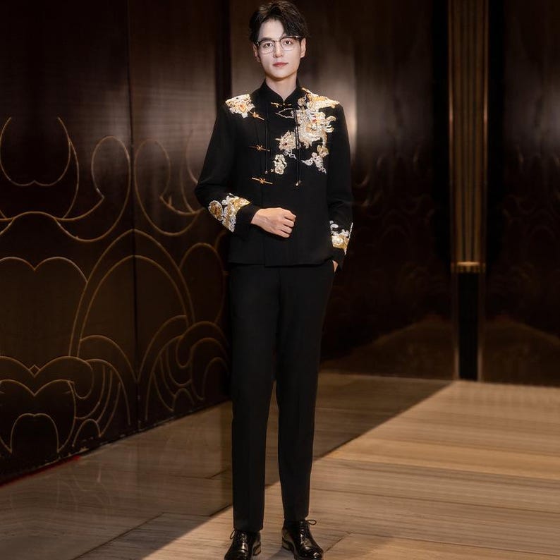 May include: A black suit with gold and white floral embroidery on the jacket. The suit includes a mandarin collar jacket and matching black pants. The jacket's sleeves and shoulders are adorned with detailed embroidery. The outfit is completed with black dress shoes.