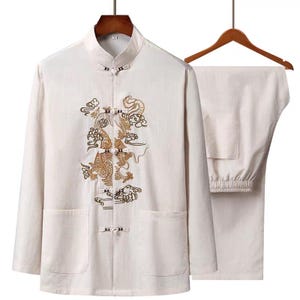 Puede incluir: Traje tradicional chino blanco roto con una parte superior de manga larga con cuello mandar&iacute;n y bordado de drag&oacute;n. Los pantalones a juego tienen cintura el&aacute;stica. El traje se exhibe en una percha de madera.