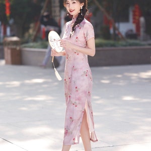 May include: A woman wearing a pink floral dress with a side slit and white shoes. She is holding a white fan with a floral design.