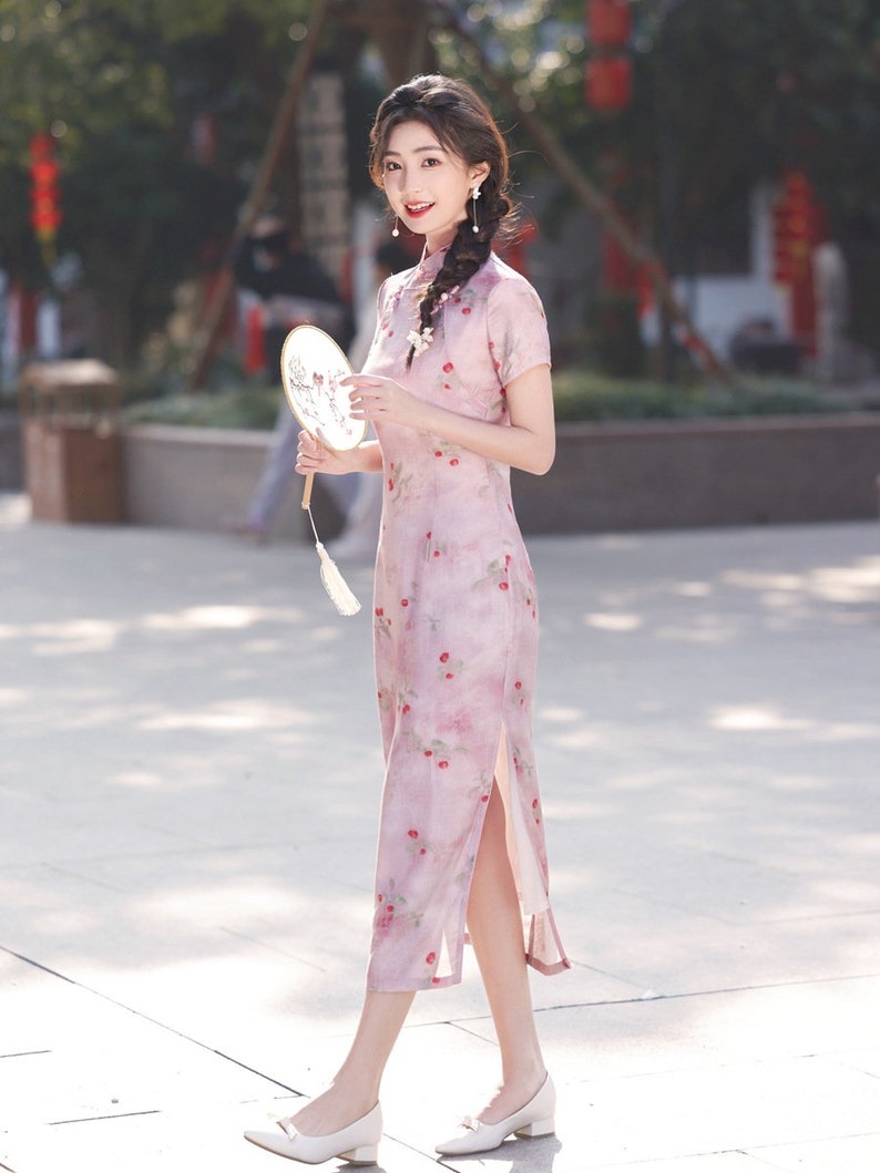 May include: A woman wearing a pink floral dress with a side slit and white shoes. She is holding a white fan with a floral design.