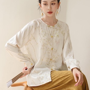 May include: A cream-colored blouse with long sleeves and a mandarin collar, adorned with a bamboo leaf pattern. The blouse features gold-colored buttons and is paired with mustard-yellow pants. The outfit is part of a traditional Asian-inspired clothing collection.