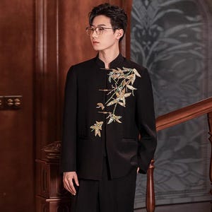 May include: Black suit with a mandarin collar jacket. The jacket features an embroidered floral design in gold, green, and white. The suit is paired with black trousers. The background includes a wooden staircase and paneling.