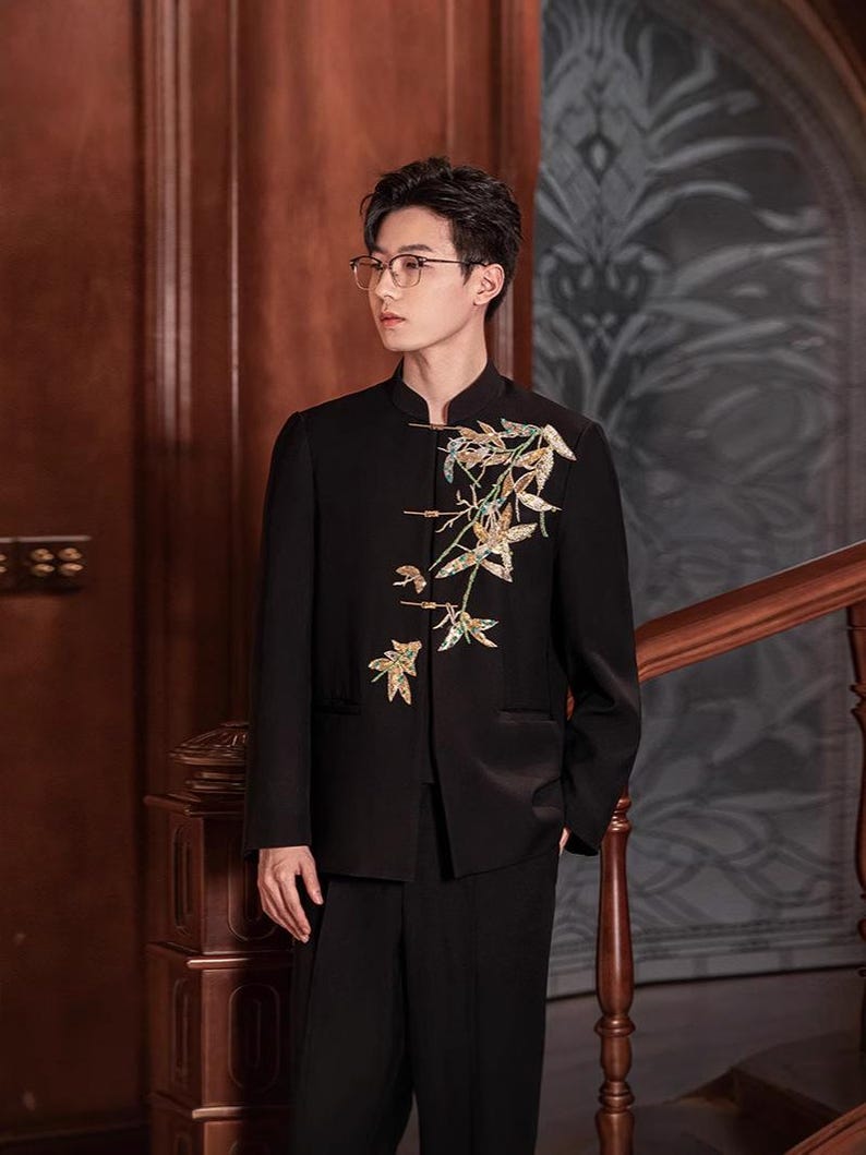 May include: Black suit with a mandarin collar jacket. The jacket features an embroidered floral design in gold, green, and white. The suit is paired with black trousers. The background includes a wooden staircase and paneling.