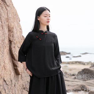 Può includere: Un top nero a maniche lunghe con colletto alla coreana e chiusure a ranella rosse. Il top ha un aspetto strutturato ed è abbinato a una gonna nera. L'outfit è indossato all'aperto vicino a una costa rocciosa.