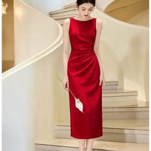 May include: A sleeveless, calf-length red dress with a boat neckline and a fitted bodice. The skirt features soft, gathered pleats along the left side, creating a draped effect. The fabric has a smooth, satin-like finish with a subtle sheen.