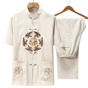 Puede incluir: Un conjunto de dos piezas en color beige claro. La parte superior es una camisa de manga corta con cuello mandarín y cierre de botones. La parte inferior es un pantalón con cintura de cordón. Ambas piezas presentan bordados intrincados en marrón y dorado.