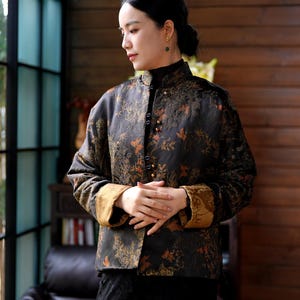 May include: A black jacket with a floral pattern in shades of gold and orange. The jacket has a mandarin collar and gold trim on the cuffs. It is buttoned up the front and appears to be made of a textured fabric.