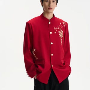 Puede incluir: Chaqueta roja brillante con cuello mandarín y bordado floral dorado en el hombro y la manga. La chaqueta tiene botones dorados y un corte entallado. La persona lleva pantalones negros. Adecuada para eventos formales.