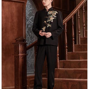 May include: A black suit with a mandarin collar jacket featuring gold floral embroidery. The suit includes matching black trousers. The person is standing on a wooden staircase with a dark brown handrail.