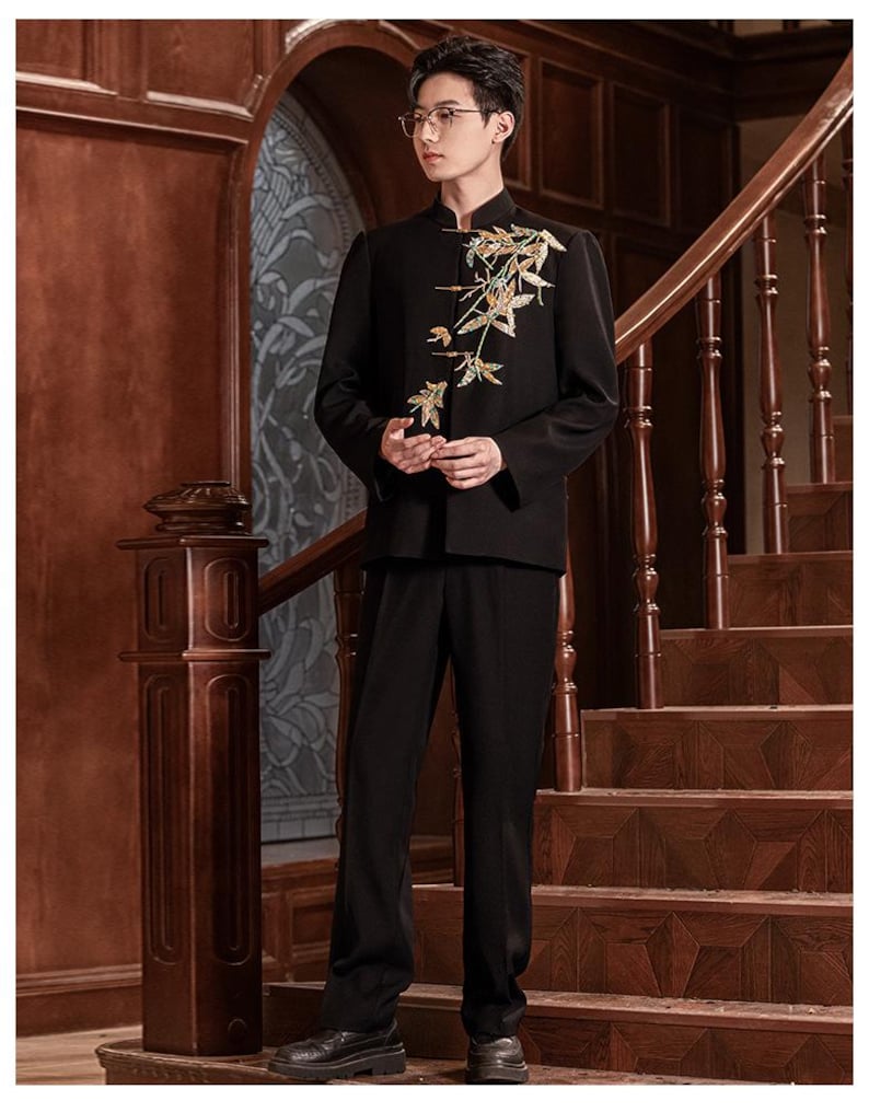 May include: A black suit with a mandarin collar jacket featuring gold floral embroidery. The suit includes matching black trousers. The person is standing on a wooden staircase with a dark brown handrail.