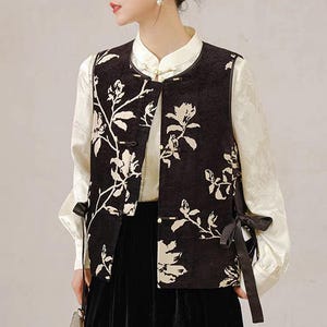 May include: A black vest with a white floral pattern. The vest has a round collar and is buttoned down the front. The sleeves are long and white. The vest is tied at the sides with black ribbons. The outfit is a traditional Asian style.
