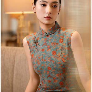 May include: A sleeveless teal dress with a mandarin collar and an orange floral pattern. The dress has a frog closure at the neckline. The model is sitting on a beige sofa.