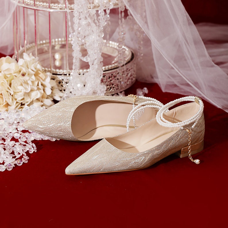 May include: A pair of gold sparkly bridal shoes with a pointed toe and a low heel. The shoes have a pearl and chain ankle strap.