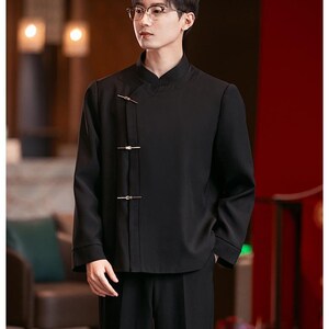 May include: Black mandarin collar shirt with gold-colored clasps. The shirt has long sleeves and is paired with black pants. The outfit has a traditional Asian-inspired design.