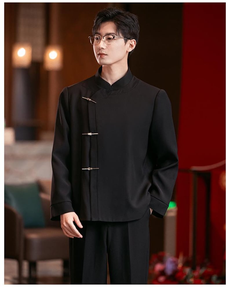 May include: Black mandarin collar shirt with gold-colored clasps. The shirt has long sleeves and is paired with black pants. The outfit has a traditional Asian-inspired design.