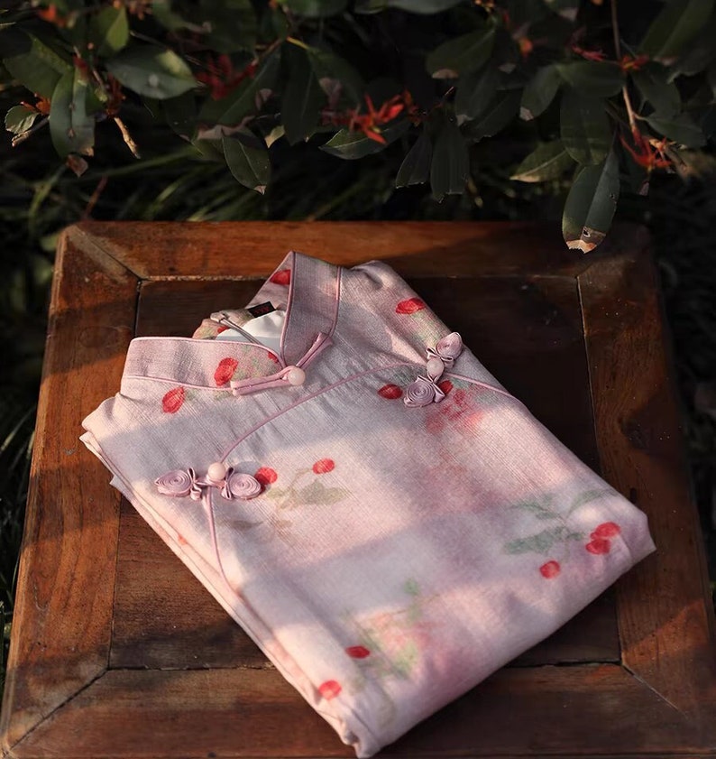 May include: A pink and white floral print dress with a mandarin collar and decorative buttons. The dress is folded and laid on a wooden surface.