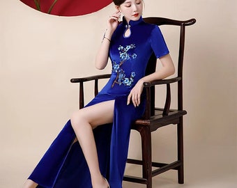 May include: A royal blue, short-sleeved dress with a high slit and floral embroidery. The dress features a mandarin collar and is made of a velvet-like material. The model is seated on a dark wooden chair.