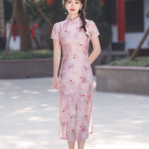 May include: A woman wearing a pink and white floral print cheongsam dress with a mandarin collar and short sleeves. She is standing on a light gray tiled surface.